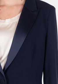 Blazer blu navy con rever in satin, vestibilità su misura, chiusura a singolo bottone e tessuto liscio, indossato su una camicia chiara.