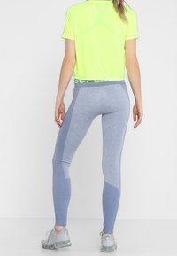 Neon zielony skrócony t-shirt, niebieskie legginsy z teksturowanymi detalami i jasnoszarymi akcentami, w zestawie z sportowymi szarymi butami.