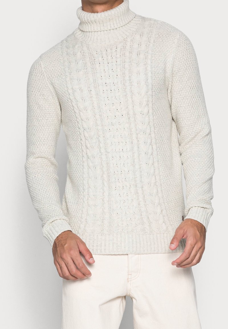 Kabelstickad polotröja i ljus gräddfärg, med texturerade mönster och ribbstickade ärmslut och nederkant, tillverkad av ett mjukt stickat material.