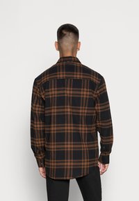 Camicia di flanella a quadri neri e arancioni con maniche lunghe, polsini abbottonati, colletto a punta e orlo arrotondato, mostrata da dietro.