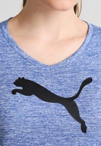 Primer plano de una persona que lleva una camiseta azul de cuello en V con una gran silueta negra de un puma saltando en el pecho.