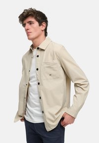 Beige knappskjorte i lett stoff med brystlomme, svarte knapper, og en hvit t-skjorte med rund hals under.