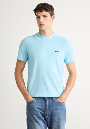 Jeune homme portant une chemise à manches courtes bleu clair avec le logo "BOSS" et un jean bleu, debout les mains dans les poches devant un fond uni.