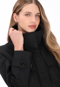 Sort jakke med høj krave, knaplukning ved manchetterne og forlommer, med en diskret tekstur. Modellen holder om kraven og fremhæver dens design.