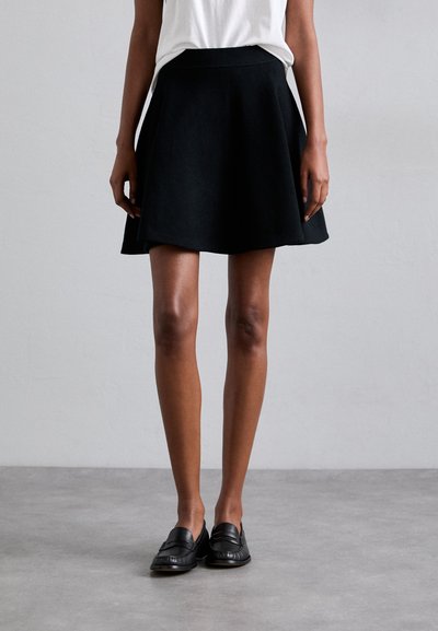 Jupe évasée noire avec une texture lisse et une taille, associée à des mocassins noirs. La tenue a un design simple et minimaliste.
