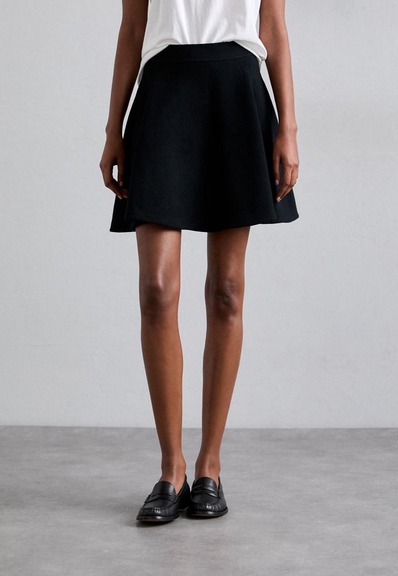 Jupe évasée noire avec une texture lisse et une taille, associée à des mocassins noirs. La tenue a un design simple et minimaliste.