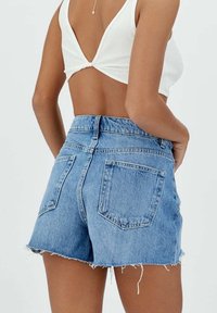 Haalarit sinisestä kulutettu farkkukangasta, joissa on revitty helma, kaksi takataskua ja klassinen viiden taskun muotoilu. Yhdistetty valkoisen ribbipaitan kanssa.