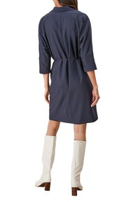 Marineblauwe jurk met riem, een kraag en driekwartmouwen, van een gestructureerde stof, gecombineerd met kniehoge witte laarzen met houten hakken.