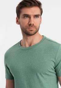 Camiseta verde de manga corta hecha de tela suave con escote redondo, que presenta una textura moteada y sutiles acentos amarillos en toda la prenda.