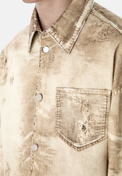 Chemise en denim beige délavée avec boutons-pression en métal, col pointu, et une poche poitrine avant avec des détails usés et effilochés.