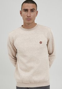 Beige sweatshirt med lange ærmer, rund hals og et lille brunt cirkulært logo på brystet. Blødt, tekstureret stof med en afslappet pasform.