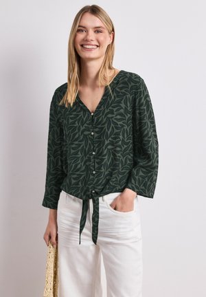 Uśmiechnięta kobieta z długimi blond włosami, ubrana w ciemnozieloną, zapinaną na guziki bluzkę z liściastym wzorem oraz białe spodnie, trzymająca plecioną torbę.