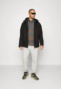 Czarna kurtka z kapturem, niebiesko-brązowa koszula w kratę, szare spodnie dresowe oraz zielone trampki z niebieskimi akcentami. Całość dopełniają okulary przeciwsłoneczne.