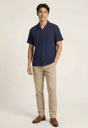 Uomo in piedi, indossa una camicia blu navy a maniche corte con bottoni, pantaloni beige e mocassini marroni, sorride con una mano in tasca.