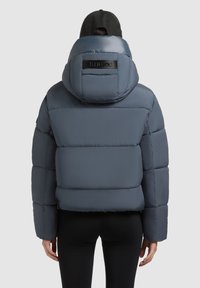 Blauwe pufferjack met capuchon, met een gequilt ontwerp, een gladde textuur en een logo-accent op de rug. Model draagt zwarte broek.