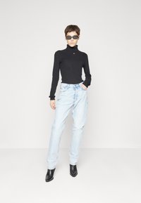 Maglione girocollo a coste nero, jeans a vita alta azzurri chiari e stivali neri con punta. Modella indossa occhiali da sole scuri. Sfondo semplice.