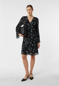 Frau in einem schwarzen Blumenkleid mit langen Ärmeln bis zum Knie, die eine schwarze Handtasche hält und schwarze spitze Absätze trägt.