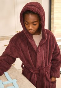 Accappatoio con cappuccio color marrone scuro, realizzato in tessuto morbido e strutturato, con cintura annodata. Chi lo indossa è in piedi davanti a un piano di lavoro con tessere da domino blu disposte sopra.