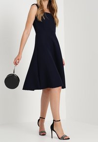 Granatowa sukienka w kształcie litery A z teksturowanej tkaniny, bez rękawów, dopasowana w talii, w połączeniu z czarnymi szpilkami na kostkę i okrągłą torebką.