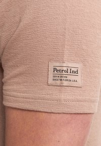 Béžová košile s krátkým rukávem z texturované látky. Na rukávu je přišitá nálepka s názvem "Petrol Ind" a podrobnostmi o značce.