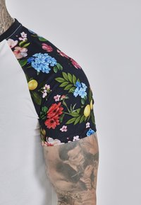 White t-shirt with black floral-patterned sleeves featuring blue, pink, and red flowers, green leaves, and yellow accents.