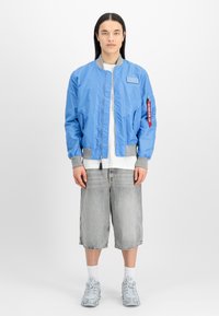 Svetlo modra bomber jakna z sivimi detajli, zadrgo, stranskimi žepi in rdečo zadrgo na poteg. V kombinaciji s sivimi kratkimi hlačami in vzorčastimi čevlji.