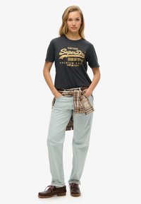 Donkergrijze t-shirt met goud vintage tekst, lichtblauwe broek met een bruine riem en een geruit overhemd geknoopt om de taille. Bruine schoenen met veters.