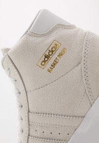 Béžové semišové tenisky s texturovaným svrškem, bílými tkaničkami a hladkou patou. Zlato-barvené logo "adidas BASKETPROOF" po straně.