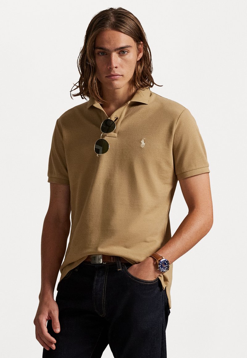 Κανώντας μια τέντα πολό, με υφασμάτινη επίστρωση, γιακά και κεντήμενο λόγο. Φόρεκε με σκόνι τζιν και γύαλια ηλίου, χέρια στις τσέπες.