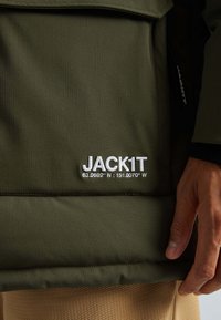 Tmavozelená bunda s bílým logem "JACK1T" a geografickými souřadnicemi na přední straně; ruka položená vedle bundy a viditelné béžové kalhoty.
