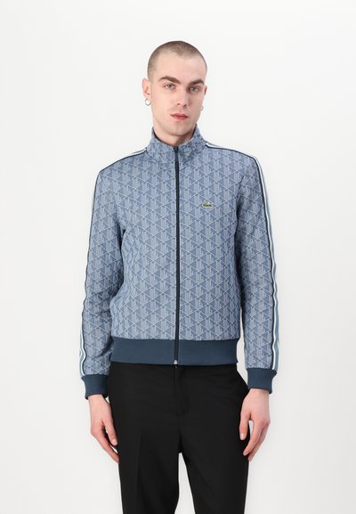 Veste zippée à motifs bleus comportant un col montant, des rayures contrastantes sur les manches et des bords côtelés aux poignets et à l'ourlet ; accent logo vert sur la poitrine.