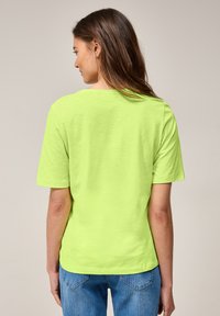 T-shirt à manches courtes et col rond de couleur vert vif, fabriqué en tissu doux. La vue de dos montre une coupe décontractée et un minimalisme dans le design. Assorti avec un jean bleu.