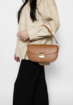 Donna con giacca beige e gonna nera tiene una borsa a mano in pelle marrone medio con chiusura dorata e tracolla.