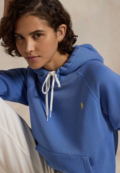 Jaunas asmuo trumpais garbanotais plaukais, dėvintis mėlynį džemperį su baltais raišteliais ir šviesias kelnes, žiūrintis tiesiai prieš save.