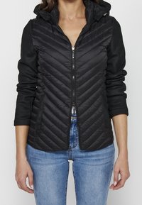 Femme portant une veste matelassée noire zippée avec capuche et manches noires, associée à un jean bleu clair.