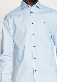 Mężczyzna w jasnoniebieskiej koszuli z długim rękawem zapinanej na guziki z małym logo Tommy Hilfiger na lewej piersi.