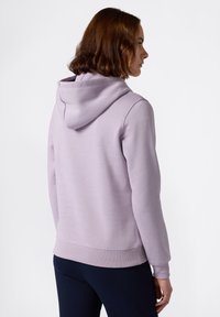 Giovane con capelli corti castani che indossa una felpa viola chiaro e pantaloni scuri, rivolto a destra su uno sfondo bianco semplice.