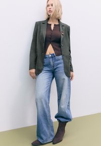 Oliven grøn blazer over en brun ribbet skjorte med knapper, parret med lyseblå vidde jeans og brune suedeboots.