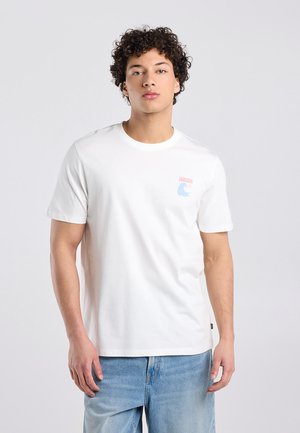 Junger Mann mit lockigem Haar, der ein weißes kurzärmeliges T-Shirt mit kleinem Logo auf der Brust und hellblaue Jeans trägt und vor einem einfarbigen Hintergrund steht.