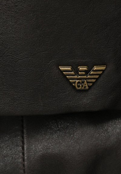 Καφέ δέρμα με υφή, διακοσμημένο με ένα μεταλλικό χρυσό έμβλημα με φτερά που έχει το "GA" στο κέντρο. Ορα τές ραφές.