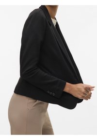 Zwarte blazer met een gestructureerd ontwerp, voorzien van een sjaalkraag, twee steekzakken aan de voorkant en manchetten met knopen. Gedragen over beige broek.