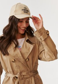 Beige trench coat doodle mintázattal és arany gombokkal, grafikus póló felett viselve. Egy bézs sapka, rajta "MY FOREVER DATE" fekete hímzéssel.