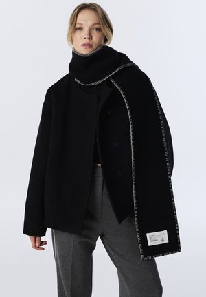 Manteau en laine noir avec un large col, associé à une écharpe noire assortie avec des coutures blanches contrastantes et une étiquette en tissu sur l'écharpe.