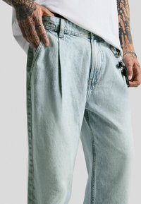 Lichtblauwe denim broek met een ontspannen pasvorm, voorzien van plooien bij de taille en zijzakken. Decoratieve hardware bevestigd aan een riemlus.