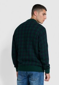 Gehaakte cardigan in een donkergroen en marineblauw patroon, met een ronde hals en geribde manchetten; de achteraanzicht benadrukt de textuur en het ontwerp.