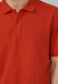 Rotes Polohemd aus strukturiertem Stoff, mit Kragen, zwei Knöpfen und einem kleinen gestickten "s.Oliver"-Logo auf der Brust.