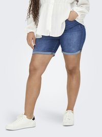 Person som bär uppvikta blå denimshorts, vit långärmad blus och vita sneakers, stående med ena handen i fickan.