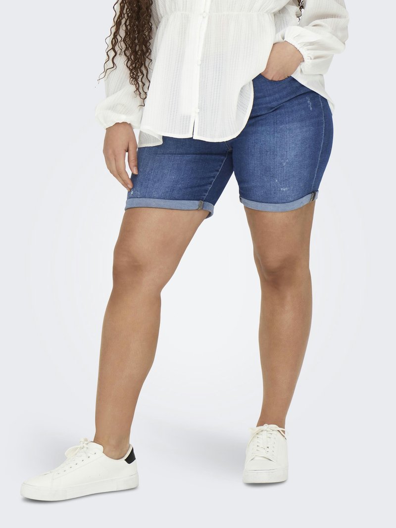 Person som bär uppvikta blå denimshorts, vit långärmad blus och vita sneakers, stående med ena handen i fickan.