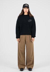 Frau in einem schwarzen Pullover, braunen weiten Hosen, schwarzen Schuhen und einer schwarzen Kappe, die vor einem schlichten hellen Hintergrund steht.