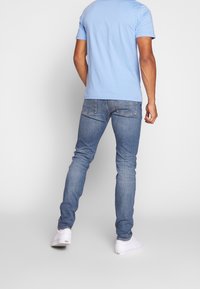 Homme portant un polo bleu clair, un jean slim-fit bleu et des baskets blanches, se tenant contre un fond blanc uni, de dos face au spectateur.